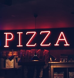 A glowing neon sign spelling 'PIZZA' dominates the scene, mounted on a dark wall above shelves that hold various items, including jars and hanging garlic. Dim lighting creates a cozy, intimate atmosphere.