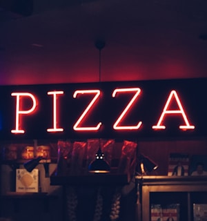 A glowing neon sign spelling 'PIZZA' dominates the scene, mounted on a dark wall above shelves that hold various items, including jars and hanging garlic. Dim lighting creates a cozy, intimate atmosphere.