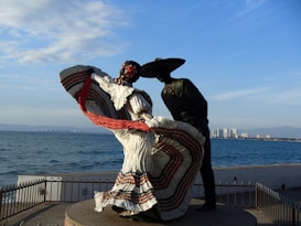 A bronze sculpture of two figures in traditional Mexican dance attire is situated near the ocean. The female dancer is wearing a flowing dress adorned with colorful stripes and a red ribbon, while the male dancer is depicted wearing a hat. The background consists of a view of the sea and a distant city skyline, with clear blue skies overhead.