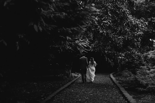 A cinematic candid of a couple walking hand in hand along a sandy path with charcoal shadows.
