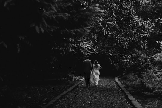 A couple is walking hand in hand down a tree-lined path. The image is in black and white, and the scene is captured from behind, emphasizing the shadows cast by the dense foliage above. The bride is wearing a long dress, and the groom is in a suit. The path is bordered by stones and covered with leaves.