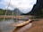 Wooden boats are resting on a calm river with towering mountains in the background. The riverbank is sandy, and the landscape is lush with greenery. The sky is partly cloudy, casting a serene atmosphere over the scene.