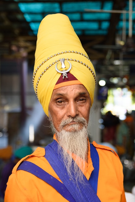 A man with a long white beard is wearing a bright yellow turban adorned with a circular metallic ornament and small beads. His outfit consists of an orange garment with blue sash details. The background is slightly blurred with hints of people and structures, suggesting a busy or crowded setting.