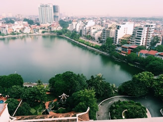 An aerial view of a city with a large lake in the center, surrounded by green trees and vegetation. Modern high-rise buildings and residential houses with red roofs line the lake's edge. There is a road running parallel to the lake. The foreground shows lush green gardens with a pathway.