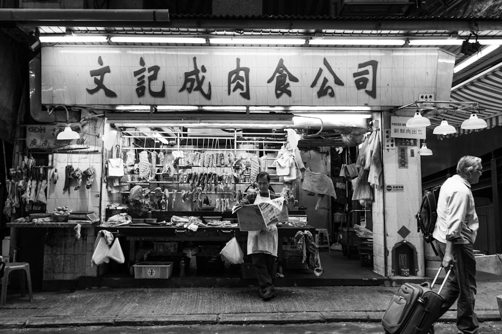 grayscale photo of person walking while holding stroller in front of store