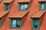 A series of dormer windows with teal frames set into a roof covered with terracotta-colored tiles. The windows are symmetrically arranged, and the overall architecture suggests a traditional style. The sunlight casts soft shadows, adding depth to the textured roof tiles.
