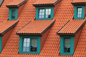 A series of dormer windows with teal frames set into a roof covered with terracotta-colored tiles. The windows are symmetrically arranged, and the overall architecture suggests a traditional style. The sunlight casts soft shadows, adding depth to the textured roof tiles.