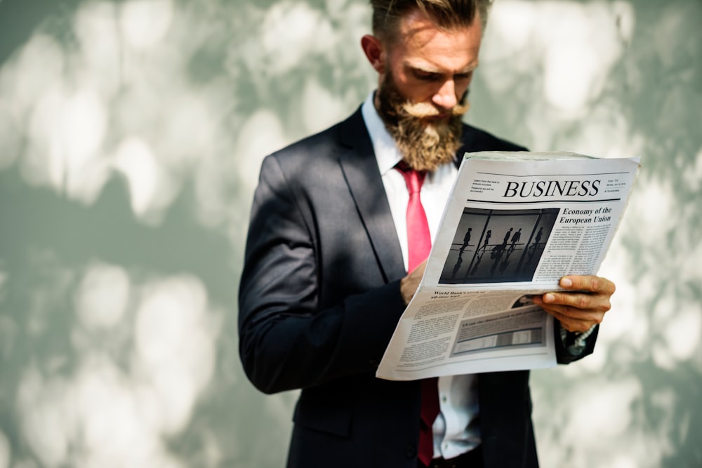 A bearded man in a suit reading the business section of a newspaper.