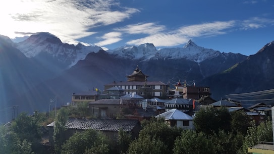 A serene mountainous landscape with a village nestled at the base of the snow-capped peaks. Traditional buildings with sloped roofs are visible, surrounded by lush greenery. The sky is clear with a few clouds, and the sunlight casts a warm glow over the scene.