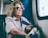 An elderly woman with curly hair and wearing sunglasses is sitting inside a train near a window. She appears thoughtful and is holding a tote bag with various designs on it. The light from the window casts a soft glow on her, highlighting her facial features.