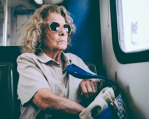 An elderly woman with curly hair and wearing sunglasses is sitting inside a train near a window. She appears thoughtful and is holding a tote bag with various designs on it. The light from the window casts a soft glow on her, highlighting her facial features.
