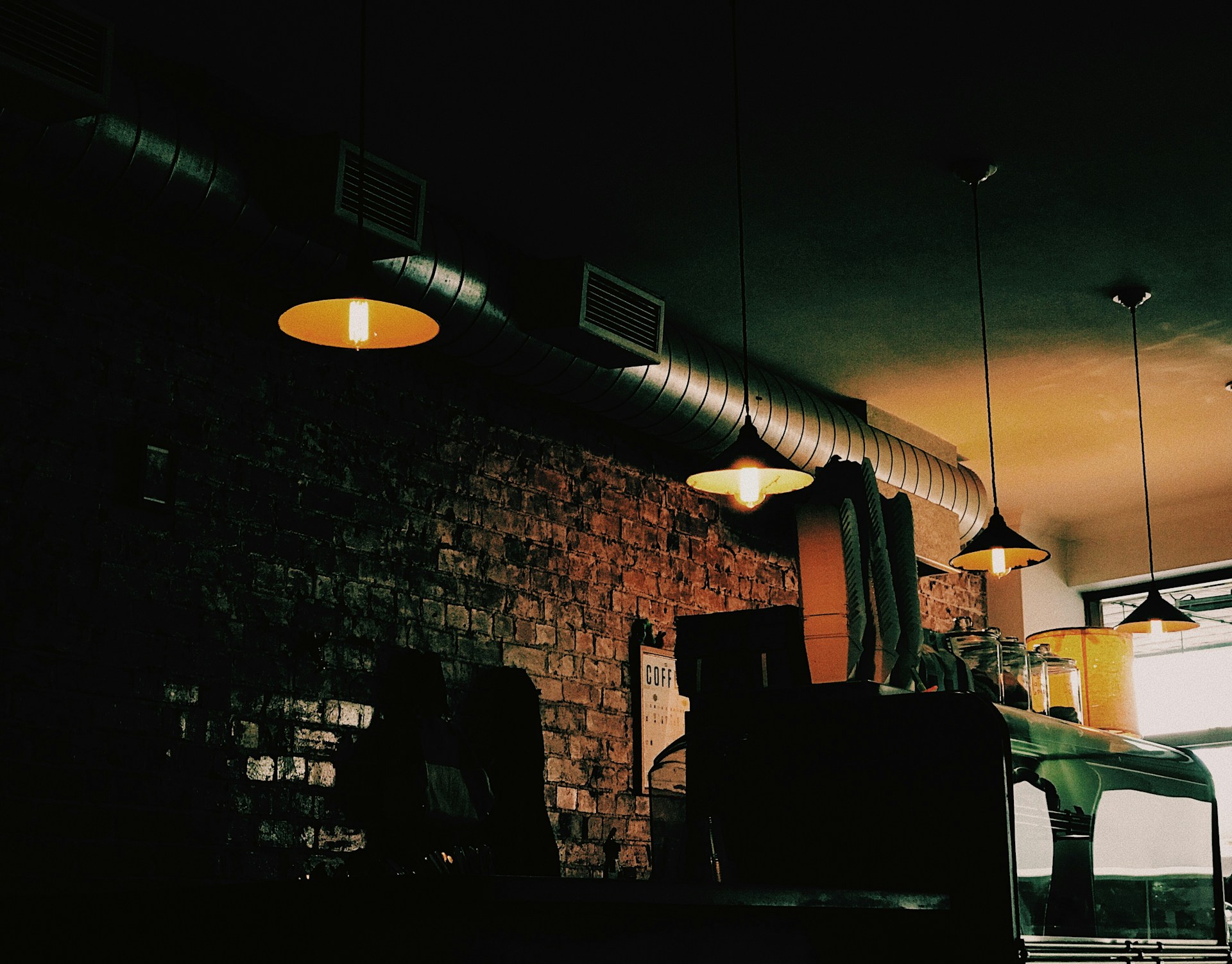 A dimly lit interior of a café with hanging lights casting a warm glow. The wall is made of exposed bricks, and there is a large metallic duct running along the ceiling. A coffee machine and various equipment are visible on the counter, along with some containers. The ambiance is cozy and slightly industrial.