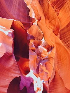 A vibrant landscape of smooth, flowing rock formations with rich shades of red and orange. Natural light filters through the crevices, highlighting the intricate textures and curves of the sandstone. Two people are visible at the bottom, emphasizing the grandeur and scale of the canyon.