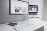 A minimalist workspace with a clean white desk featuring a desktop monitor displaying a webpage, alongside an elevated laptop showing another webpage. There is a white keyboard, a mouse, a smartphone on top of a closed notebook, and earphones neatly placed on the table.
