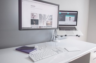 A minimalist workspace with a clean white desk featuring a desktop monitor displaying a webpage, alongside an elevated laptop showing another webpage. There is a white keyboard, a mouse, a smartphone on top of a closed notebook, and earphones neatly placed on the table.