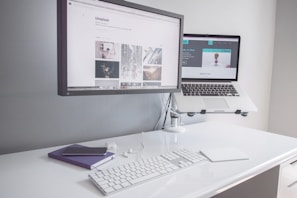 A minimalist workspace with a clean white desk featuring a desktop monitor displaying a webpage, alongside an elevated laptop showing another webpage. There is a white keyboard, a mouse, a smartphone on top of a closed notebook, and earphones neatly placed on the table.