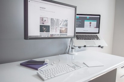A minimalist workspace with a clean white desk featuring a desktop monitor displaying a webpage, alongside an elevated laptop showing another webpage. There is a white keyboard, a mouse, a smartphone on top of a closed notebook, and earphones neatly placed on the table.