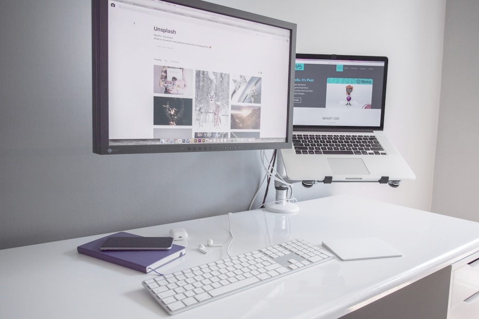 A minimalist workspace with a clean white desk featuring a desktop monitor displaying a webpage, alongside an elevated laptop showing another webpage. There is a white keyboard, a mouse, a smartphone on top of a closed notebook, and earphones neatly placed on the table.