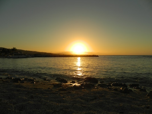 A serene beach scene with a golden sunset on the horizon, casting a warm glow on the water. The sun's reflection creates a shimmering path on the gentle waves. In the distance, silhouettes of hills and palm trees add to the tranquil setting.