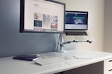 A minimalist desk setup features a desktop monitor attached to the wall and a laptop on a stand, both displaying different web pages. The desk also holds a keyboard, a mouse, a notebook, and a smartphone, creating an organized and efficient workspace.