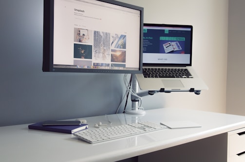 A minimalist desk setup features a desktop monitor attached to the wall and a laptop on a stand, both displaying different web pages. The desk also holds a keyboard, a mouse, a notebook, and a smartphone, creating an organized and efficient workspace.