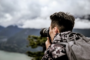 A person is taking a photograph with a camera, facing a scenic landscape with cloudy skies and distant mountains. The individual is wearing a floral-patterned jacket and carrying a gray backpack.