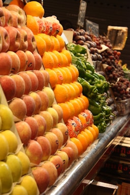 A market stall neatly displays rows of fresh fruits including peaches, oranges, and grapes, alongside green peppers. The produce is organized in sections, showcasing vibrant colors and inviting freshness.