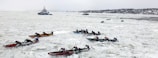 Multiple teams are participating in an ice canoe race on a frozen river, with competitors paddling through thick ice. In the background, a couple of vessels are navigating through the icy waters, while a distant shoreline is visible.