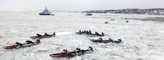 Multiple teams are participating in an ice canoe race on a frozen river, with competitors paddling through thick ice. In the background, a couple of vessels are navigating through the icy waters, while a distant shoreline is visible.