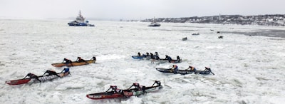 Multiple teams are participating in an ice canoe race on a frozen river, with competitors paddling through thick ice. In the background, a couple of vessels are navigating through the icy waters, while a distant shoreline is visible.