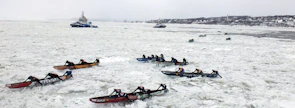 Multiple teams are participating in an ice canoe race on a frozen river, with competitors paddling through thick ice. In the background, a couple of vessels are navigating through the icy waters, while a distant shoreline is visible.