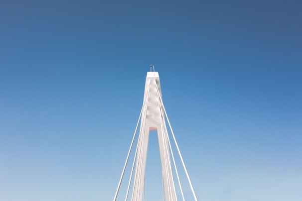 A towering cable-stayed bridge under construction, with cranes and workers visible against a clear blue sky.