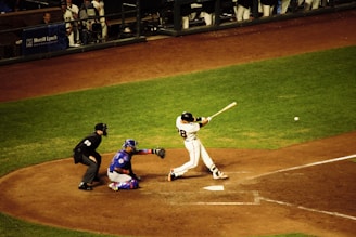 A baseball player is in mid-swing at a pitch during a game. The batter is dressed in a white uniform with the number 28 on the back, and nearby, a catcher in blue gear is crouched ready to catch the ball. An umpire in black attire watches closely. The surrounding field is a mix of green grass and brown dirt, and other team members are visible in the dugout.