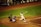 A baseball player is in mid-swing at a pitch during a game. The batter is dressed in a white uniform with the number 28 on the back, and nearby, a catcher in blue gear is crouched ready to catch the ball. An umpire in black attire watches closely. The surrounding field is a mix of green grass and brown dirt, and other team members are visible in the dugout.