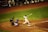 A baseball player is in mid-swing at a pitch during a game. The batter is dressed in a white uniform with the number 28 on the back, and nearby, a catcher in blue gear is crouched ready to catch the ball. An umpire in black attire watches closely. The surrounding field is a mix of green grass and brown dirt, and other team members are visible in the dugout.