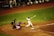 A baseball player is in mid-swing at a pitch during a game. The batter is dressed in a white uniform with the number 28 on the back, and nearby, a catcher in blue gear is crouched ready to catch the ball. An umpire in black attire watches closely. The surrounding field is a mix of green grass and brown dirt, and other team members are visible in the dugout.