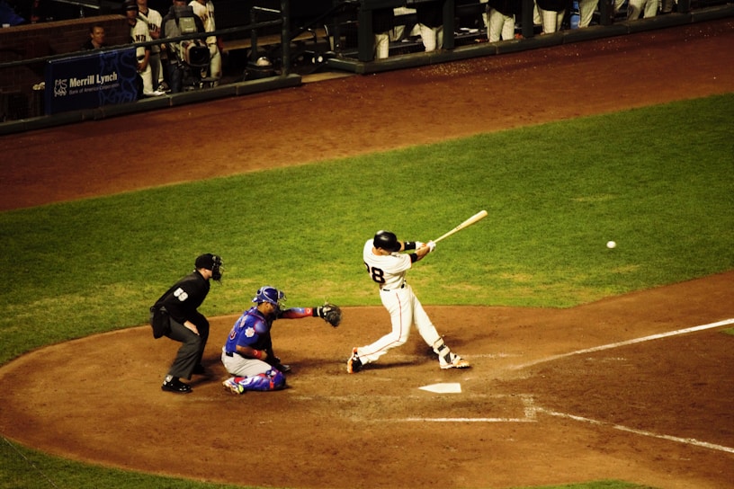 A baseball player is in mid-swing at a pitch during a game. The batter is dressed in a white uniform with the number 28 on the back, and nearby, a catcher in blue gear is crouched ready to catch the ball. An umpire in black attire watches closely. The surrounding field is a mix of green grass and brown dirt, and other team members are visible in the dugout.