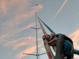 A close-up view of nautical ropes secured with knots, with a sailboat mast extending upwards against a backdrop of a sky painted with soft clouds and gentle hues.