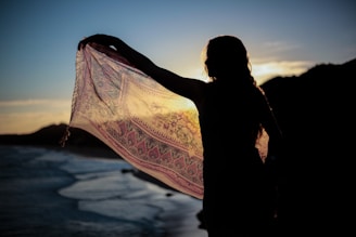 A silhouette of a person holding a patterned scarf or shawl against a sunset backdrop, with a body of water and hills in the distance. The setting sun creates a dramatic contrast, highlighting the intricate details of the fabric.