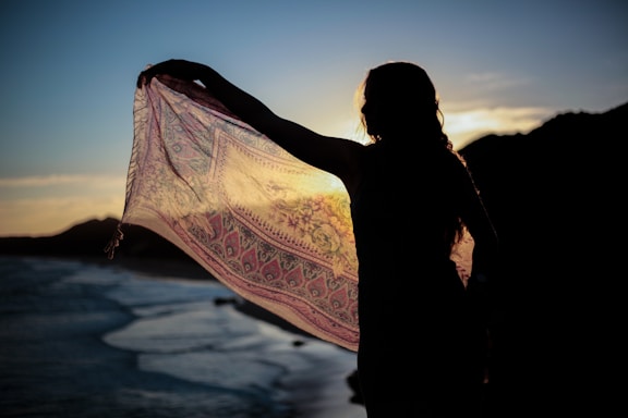 A silhouette of a person holding a patterned scarf or shawl against a sunset backdrop, with a body of water and hills in the distance. The setting sun creates a dramatic contrast, highlighting the intricate details of the fabric.