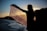 A silhouette of a person holding a patterned scarf or shawl against a sunset backdrop, with a body of water and hills in the distance. The setting sun creates a dramatic contrast, highlighting the intricate details of the fabric.