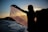 A silhouette of a person holding a patterned scarf or shawl against a sunset backdrop, with a body of water and hills in the distance. The setting sun creates a dramatic contrast, highlighting the intricate details of the fabric.