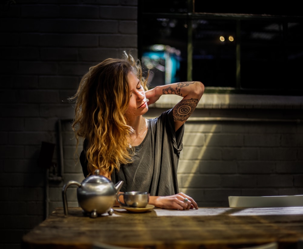 Femme en haut gris assise à côté d’une théière grise et d’une tasse sur une table en bois brun