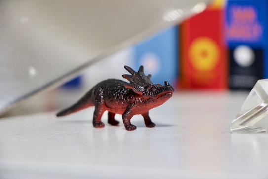 A small, toy dinosaur with a red and black color palette stands on a white surface. Its body and facial features are detailed, and it is positioned near some colorful blurred objects in the background that appear to be books or boxes. A clear, triangular object is positioned on the right side of the frame.