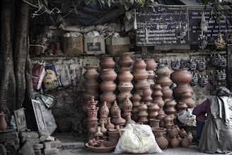 A shop or stall displays an array of terracotta pots of various sizes and designs. Some are stacked in tall columns, while others are placed on the ground. Next to the pots, a person is seated, possibly attending to the products. The background includes a mix of bags, boxes, and a sign with text in multiple languages. The setting appears rustic and is shaded by tree foliage.