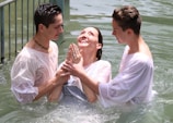 Three people are standing in a body of water, appearing to be engaged in a baptism or religious ritual. Two individuals support a third, who is in the middle, looking upwards with a joyful expression. All participants are wearing white garments, partially wet from the water.