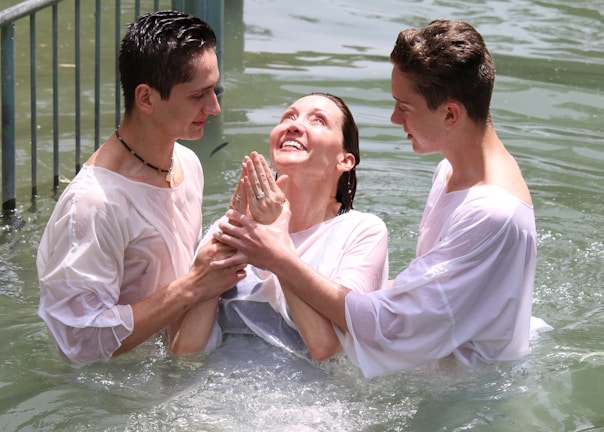 Three people are standing in a body of water, appearing to be engaged in a baptism or religious ritual. Two individuals support a third, who is in the middle, looking upwards with a joyful expression. All participants are wearing white garments, partially wet from the water.
