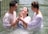 Three people are standing in a body of water, appearing to be engaged in a baptism or religious ritual. Two individuals support a third, who is in the middle, looking upwards with a joyful expression. All participants are wearing white garments, partially wet from the water.