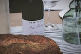 A rustic scene featuring a bottle of wine labeled 'Hewitson Miss Harry 2013 Barossa Valley' surrounded by a loaf of crusty bread and a few glassware items on a striped tablecloth. In the background, there is a green glass bottle and a clear wine glass.