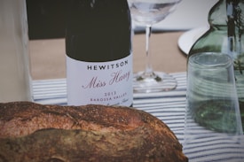 A rustic scene featuring a bottle of wine labeled 'Hewitson Miss Harry 2013 Barossa Valley' surrounded by a loaf of crusty bread and a few glassware items on a striped tablecloth. In the background, there is a green glass bottle and a clear wine glass.
